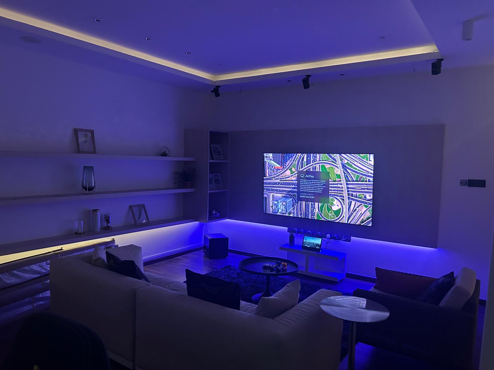 A large living room with a big screen TV, two couches, and a coffee table. The room is dimly lit, with blue lighting illum...